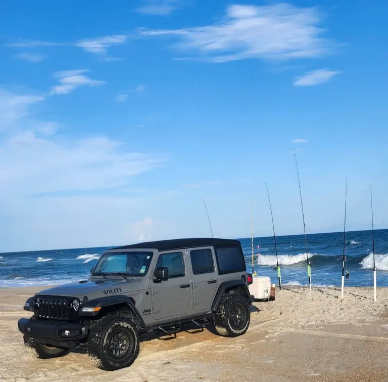 A gray Jeep Wrangler parked on the beach beside a row of surf rods in sand spikes and a cooler, with waves breaking behind