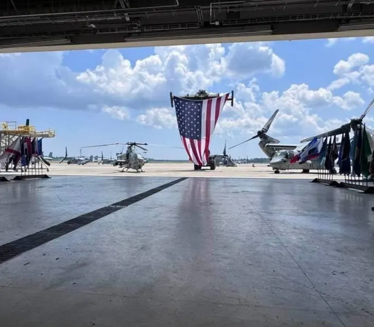 A large American flag hangs in the open door of an aviation hangar, lined with unit flags, with a Marine helicopter and a tilt-rotor aircraft on the flight line under a blue sky