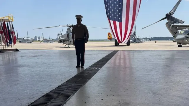 A Marine in dress uniform stands in silhouette in an open aircraft hangar, with a large American flag and military helicopters behind him, on his retirement day after more than 25 years of service